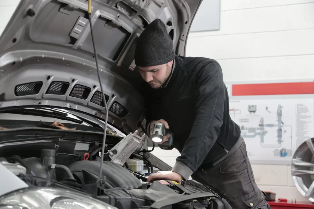 Mechanik w czapce oświetla latarką komorę silnika samochodu w warsztacie